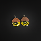 A pair of handmade earrings featuring a semi-circular wooden top with the brand name 'MAMAZZO' engraved, adorned with three dangling strands of beads in yellow, lime green, and white, set against a solid black background.