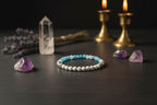 A beaded bracelet with light blue, white, and gray beads sits on a dark wooden surface, surrounded by crystals, dried lavender, a clear quartz point, and two lit brass candlesticks in the background.