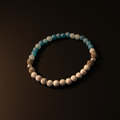 A beaded stretch bracelet arranged in a circle on a dark brown background. The bracelet features light blue and white marbled beads, white stone beads, textured brown porous beads, and small silver spacer beads.
