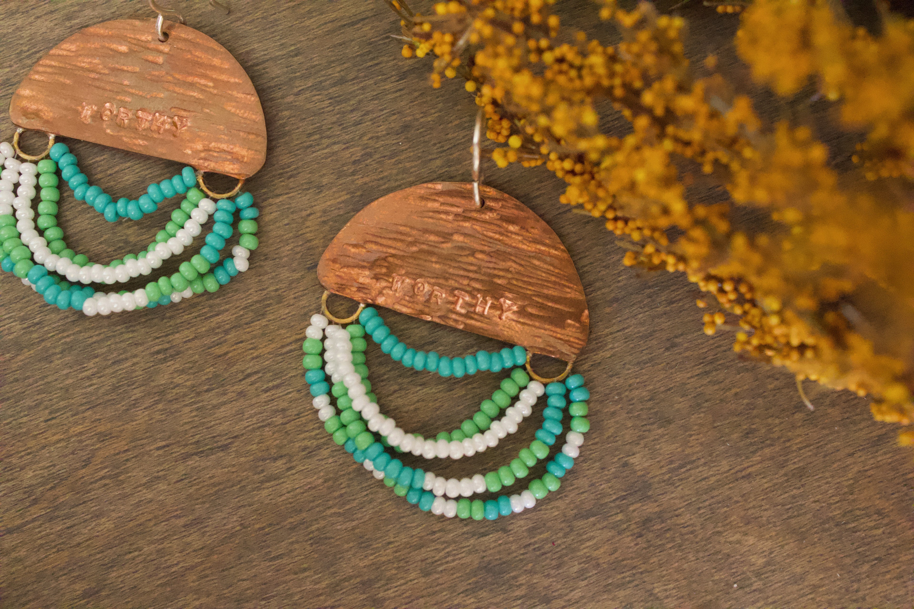 Close-up of Mini Wordkeeper Earrings Worthy by Jewellery by JN, highlighting textured copper, green, teal, and white beadwork, and hand-stamped word WORTHY.
