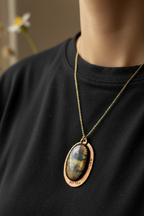 A close-up of a person wearing a gold-toned oval pendant necklace with a tiger's eye stone, styled over a black crewneck shirt.