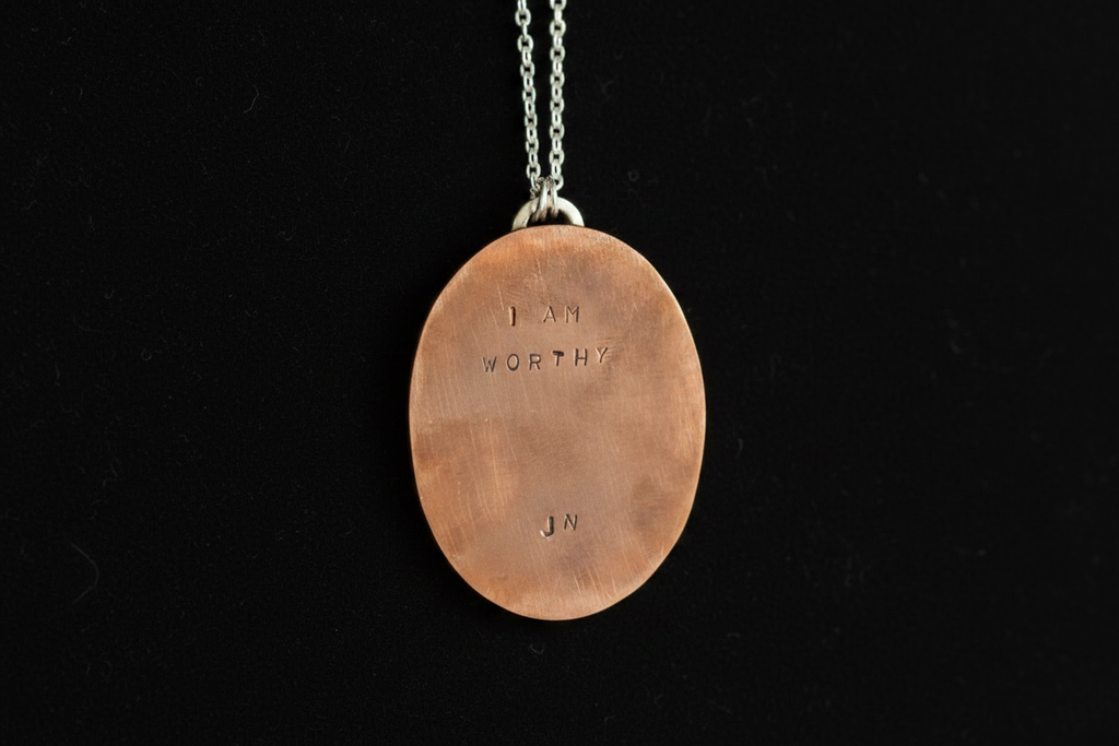 A copper oval pendant necklace engraved with 'I AM WORTHY' and 'JN', suspended from a silver chain against a black background.