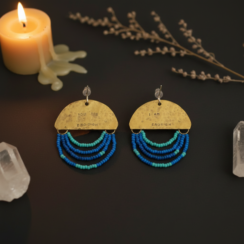 A pair of handmade statement earrings displayed on a dark surface with decorative props, including a lit candle, dried botanicals, and clear crystals. The earrings feature a textured gold-toned semi-circle top with the words 'YOU ARE ENOUGH' and 'I AM ENOUGH' printed on them, with three tiers of blue and turquoise beaded fringe below.