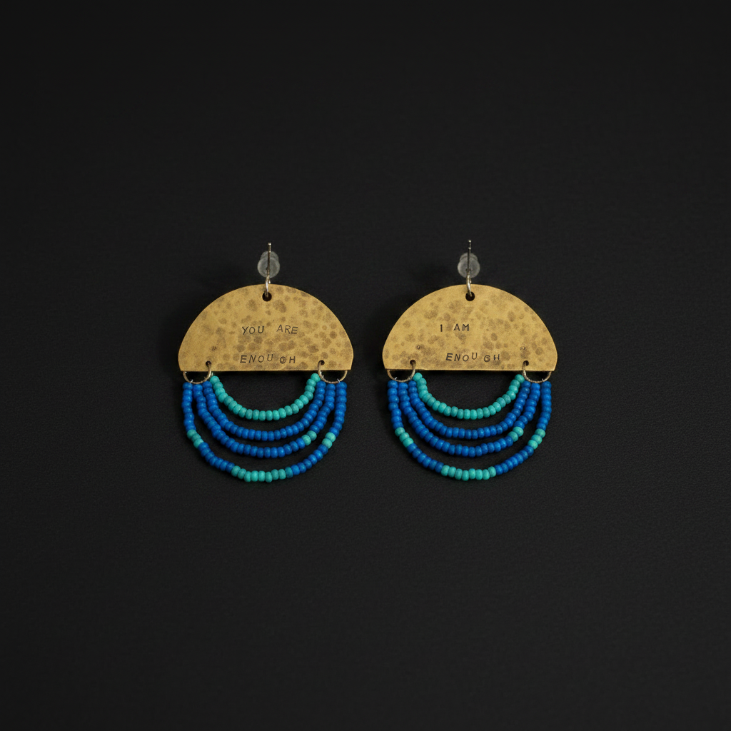 A pair of handmade dangle earrings on a solid black background. Each earring features a hammered gold-tone semicircle disc engraved with 'YOU ARE ENOUGH' and 'I AM ENOUGH', with three curved strands of small beads in shades of blue, teal, and turquoise hanging below.