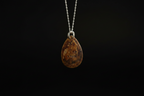 A teardrop-shaped brown gemstone pendant with natural inclusions, suspended from a silver chain against a solid black background.