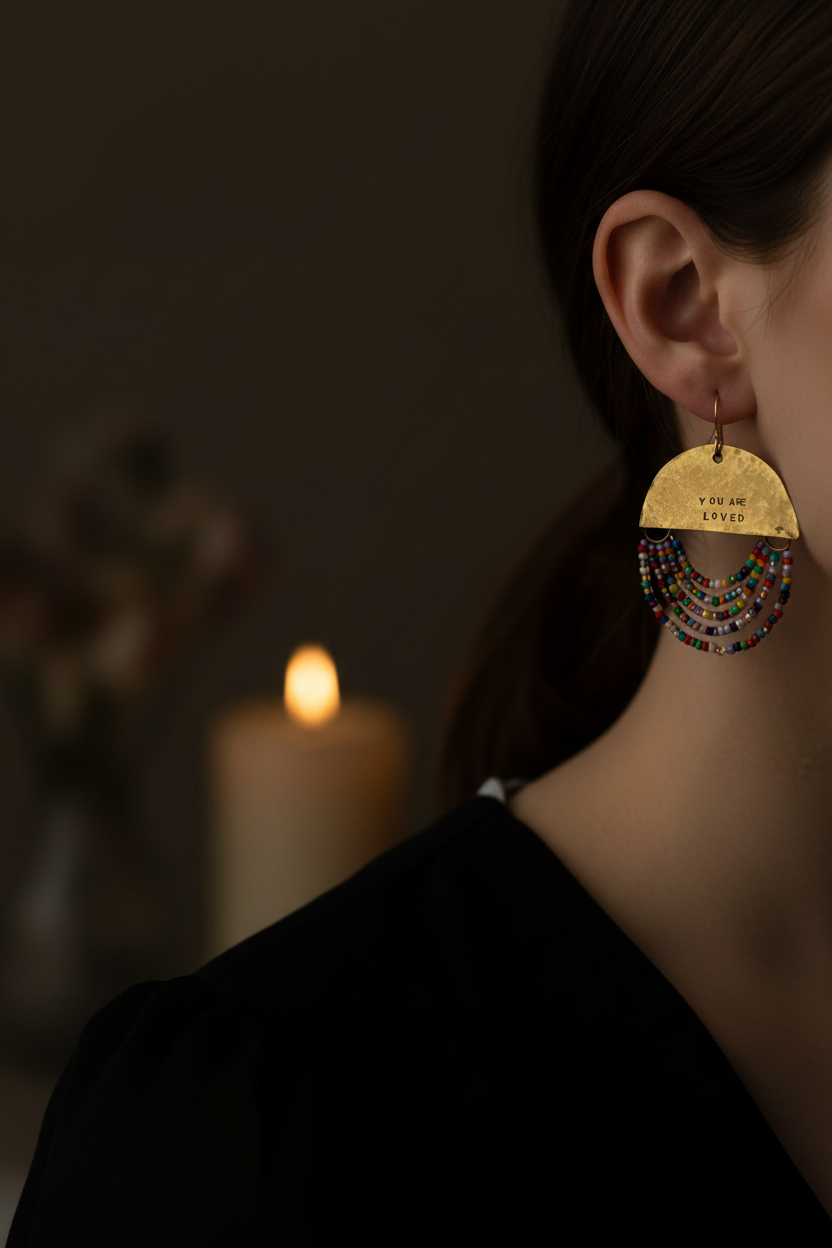 A close-up side profile of a woman wearing a large gold-toned dangle earring with engraved text and layered multicolor beaded fringe, against a dark background with a blurred candle.