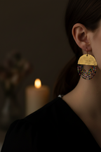A close-up side profile of a woman wearing a large gold-toned dangle earring with engraved text and layered multicolor beaded fringe, against a dark background with a blurred candle.
