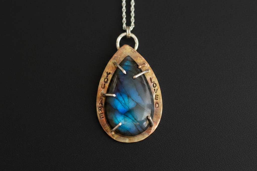 A teardrop-shaped pendant necklace featuring a blue labradorite stone with silver prongs, set in a rustic bronze-toned bezel engraved with 'LOVED' and 'YOU ARE WELCOME'.