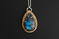A teardrop-shaped pendant necklace featuring a blue labradorite stone with silver prongs, set in a rustic bronze-toned bezel engraved with 'LOVED' and 'YOU ARE WELCOME'.