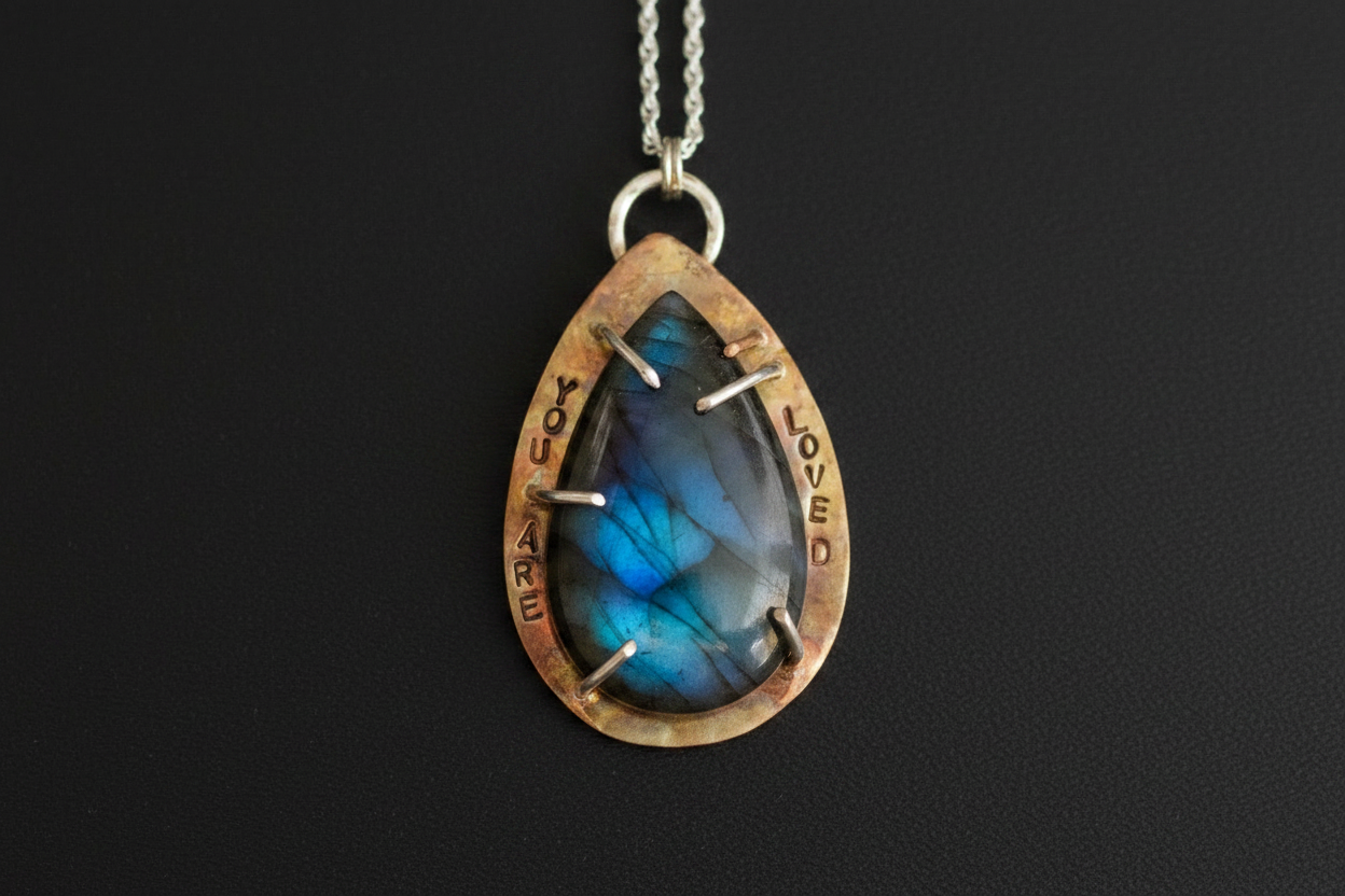 A teardrop-shaped pendant necklace featuring a blue labradorite stone with silver prongs, set in a rustic bronze-toned bezel engraved with 'LOVED' and 'YOU ARE WELCOME'.