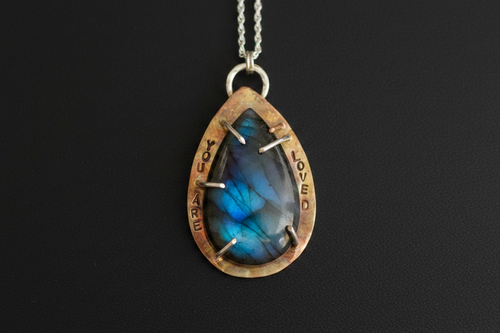 A teardrop-shaped pendant necklace featuring a blue labradorite stone with silver prongs, set in a rustic bronze-toned bezel engraved with 'LOVED' and 'YOU ARE WELCOME'.