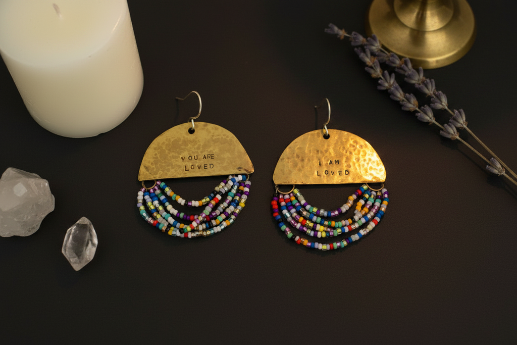 A pair of handmade gold-toned dangle earrings with beaded fringe, displayed on a dark surface with a white candle, crystals, and dried lavender. The top semi-circle of each earring is engraved with 'YOU ARE LOVED'.