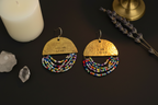 A pair of handmade gold-toned dangle earrings with beaded fringe, displayed on a dark surface with a white candle, crystals, and dried lavender. The top semi-circle of each earring is engraved with 'YOU ARE LOVED'.