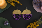 A pair of handmade dangle earrings displayed on a dark background with decorative botanical props. The earrings feature a hammered brass semi-circle top with engraved text, and multiple strands of purple and silver beads hanging below.