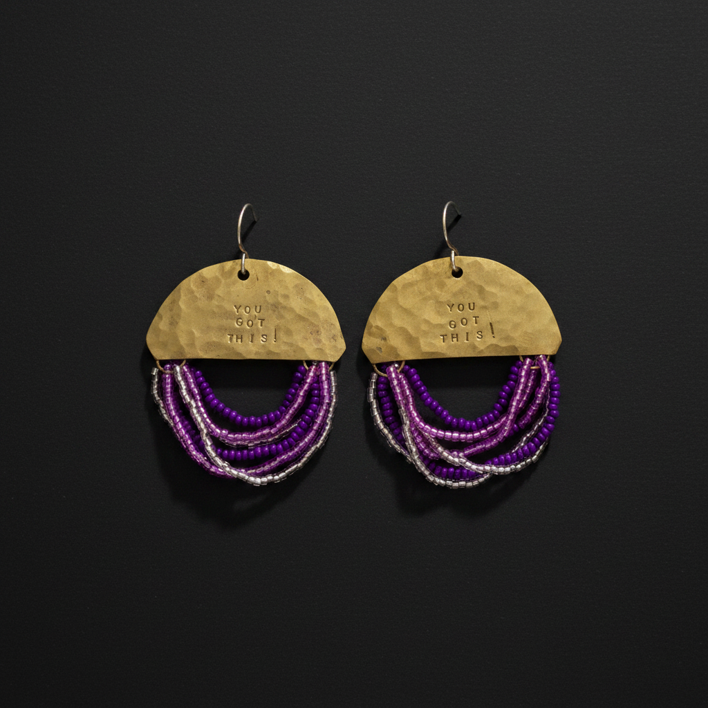 A pair of handmade dangle earrings on a dark background. Each earring features a hammered gold-tone semicircle top stamped with 'YOU GOT THIS!' and multiple strands of purple and silver beads hanging below.